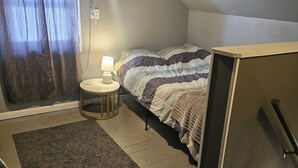 Een slaapkamer met een bed, nachtkastje, lamp, raam met gordijnen, vloerkleed en een trap.