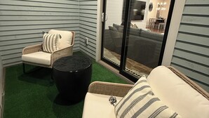 Una habitación con piso de césped artificial verde, dos sofás de mimbre con cojines a rayas, un reposapiés negro y una puerta corrediza de vidrio que da a una zona de estar contigua.