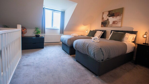 A bedroom with two beds, a radiator, a window with curtains, a plant, and a painting on the wall. - Coventry House Near City Centre & Arena (Coventry)