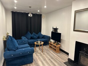 Un salon avec un canapé bleu, une petite table ronde, un téléviseur à écran plat, une cheminée et un luminaire au plafond.
