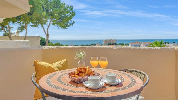 A round table with a basket of croissants, two cups, and two glasses of orange juice, set against a scenic view of the sea and trees. - Luma Riviera, relaxing with sea view and pool. Walking distance to the beach. (Mijas)