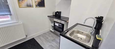 A compact kitchen with a black countertop, stainless steel sink, and a built-in oven. There are two framed abstract paintings on the wall, a window with blinds, and a radiator.