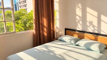 Un lit avec une tête de lit en bois, une fenêtre avec des rideaux et une vue sur la verdure à l’extérieur.