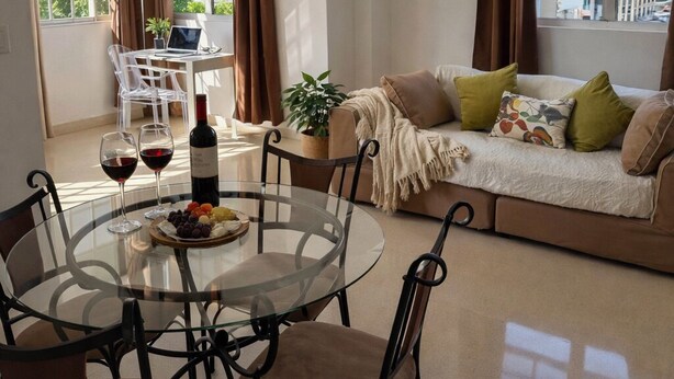 A modern living room with a glass dining table, a sofa with cushions, a potted plant, and a view of buildings through large windows. - Bright & Spacious Apartment in Obarrio | Walk to Everything + City Views (Panamá)