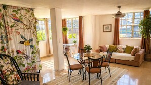 A living room with a round glass table, a sofa, and a chair. There is a window with curtains, a plant, and a framed picture on the wall. - Bright & Spacious Apartment in Obarrio | Walk to Everything + City Views (Panamá)