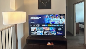 Ein Wohnzimmer mit Wandfernseher, einer Lampe, einem Hirschgeweih im Bilderrahmen und einer Treppe, die in einen anderen Raum führt.