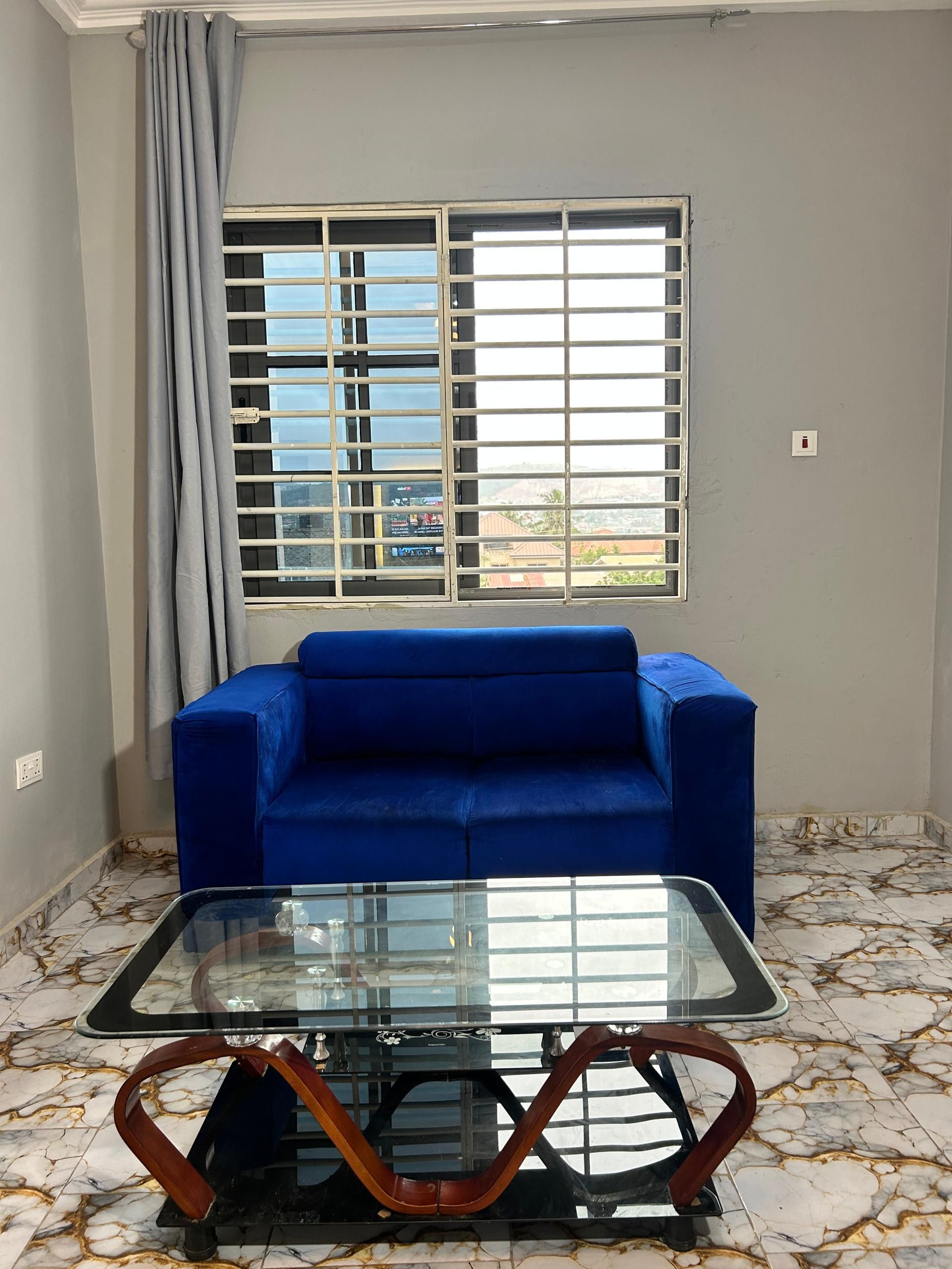 Una habitación con un sofá azul, una mesa de centro con superficie de vidrio y patas de madera, una ventana con persianas y piso de baldosas.