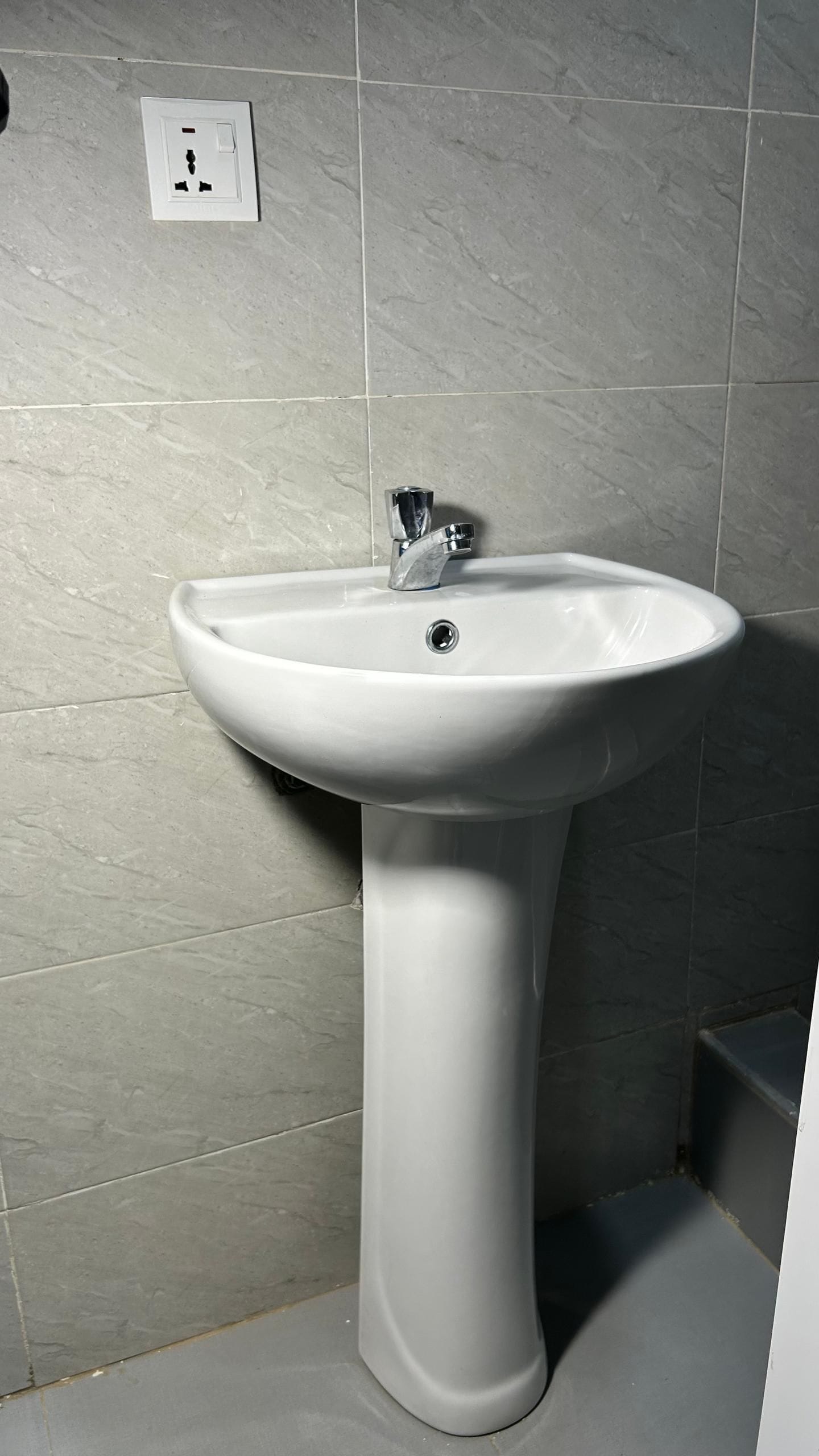 Een badkamer met een wastafel op een voetstuk, een lichtschakelaar aan de muur en een betegelde vloer.