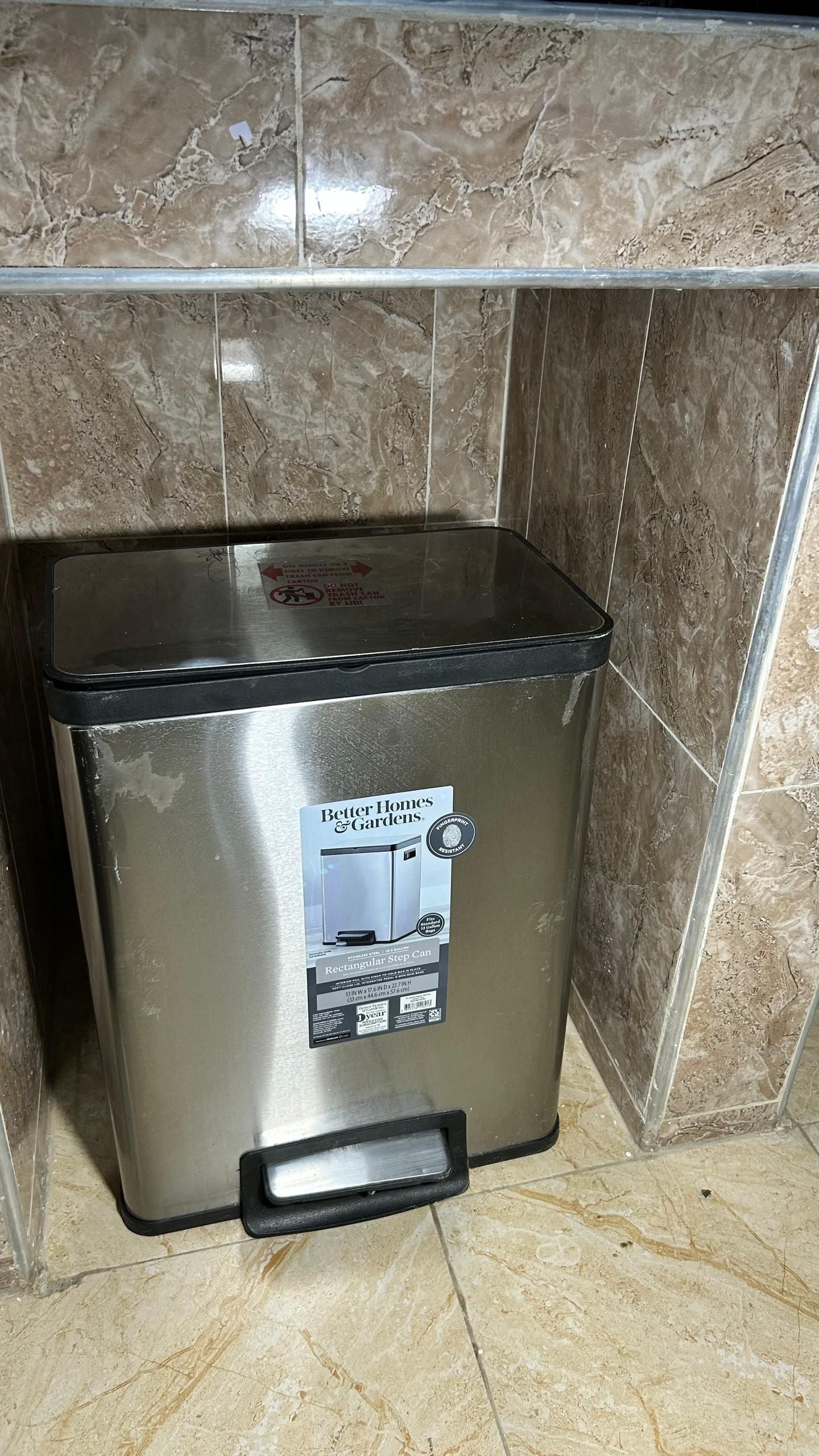 A stainless steel trash can with a lid, placed against a tiled wall.