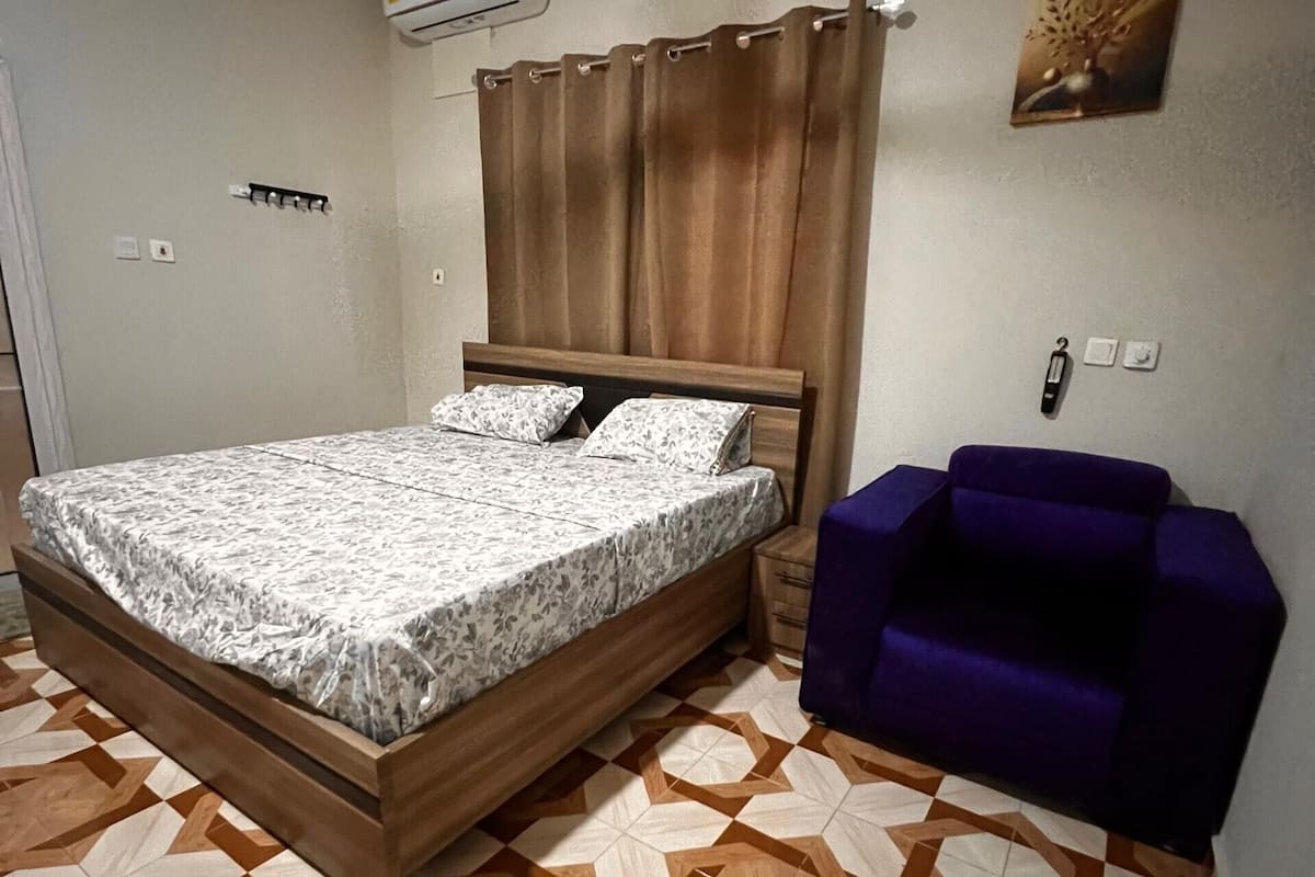 Un dormitorio con cama, aire acondicionado, sofá morado y suelo estampado.