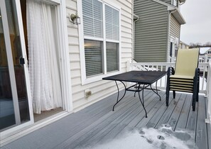 Eine Holzdielen-Terrasse mit Tisch, Stuhl, einer Glasschiebetür und einem Fenster mit Jalousien.