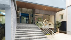 Una escalera conduce a la entrada de un edificio, con barandilla metálica, una planta en maceta y una puerta con ventana.