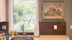 A kitchen with a sink, faucet, window with blinds, and a framed painting on the wall. - Mountain House in Town (Falls Church)