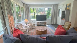 A living room with a sofa, a television, wicker chairs, and a view of greenery outside. - Mountain House in Town (Falls Church)