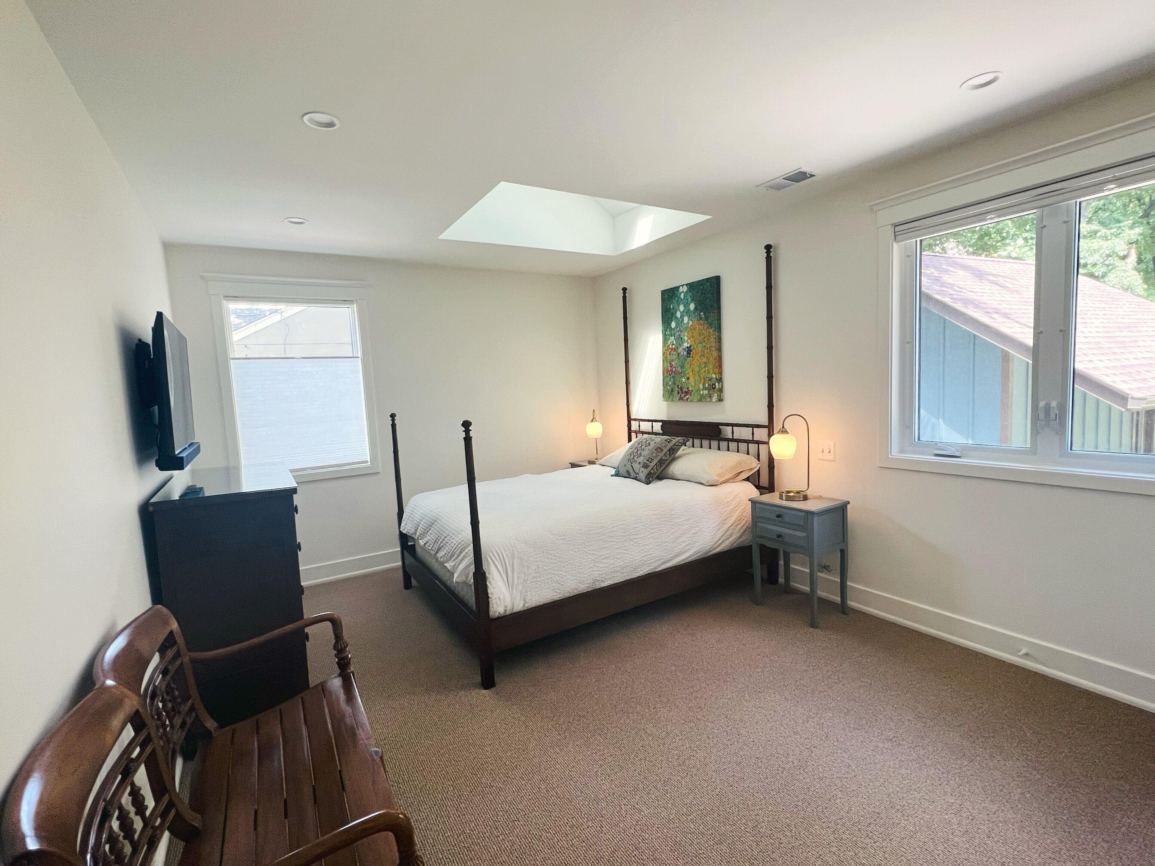 A bedroom with a bed, bedside tables, a desk, a chair, a painting, and a skylight.