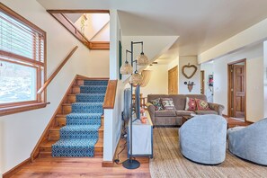 Una escalera con alfombra estampada, una sala de estar con sofá y cojines, un reposapiés redondo y un lámpara colgante.
