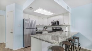 Cozinha moderna com armários brancos, geladeira de aço inoxidável, micro-ondas e ilha de granito com banquetas.