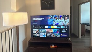 Una sala de estar con un televisor de pantalla plana montado en la pared, una lámpara, una cabeza de venado enmarcada y una escalera que lleva a otra habitación.