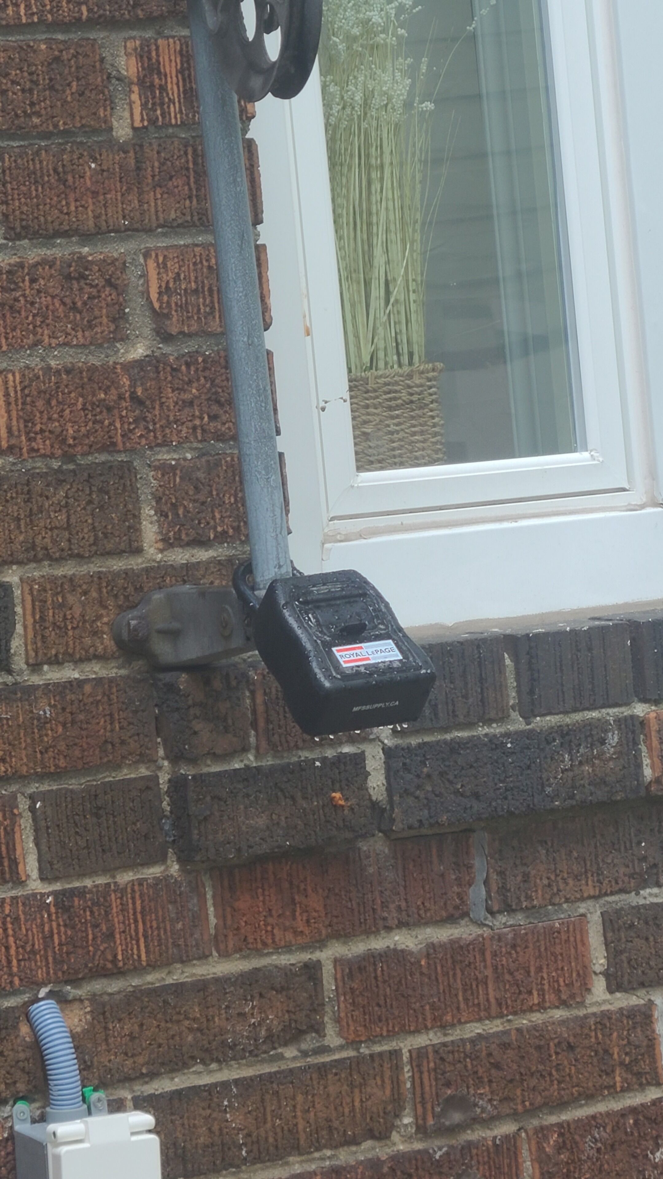 A black electrical box mounted on a brick wall, a window with a white frame, and a plant in the background.