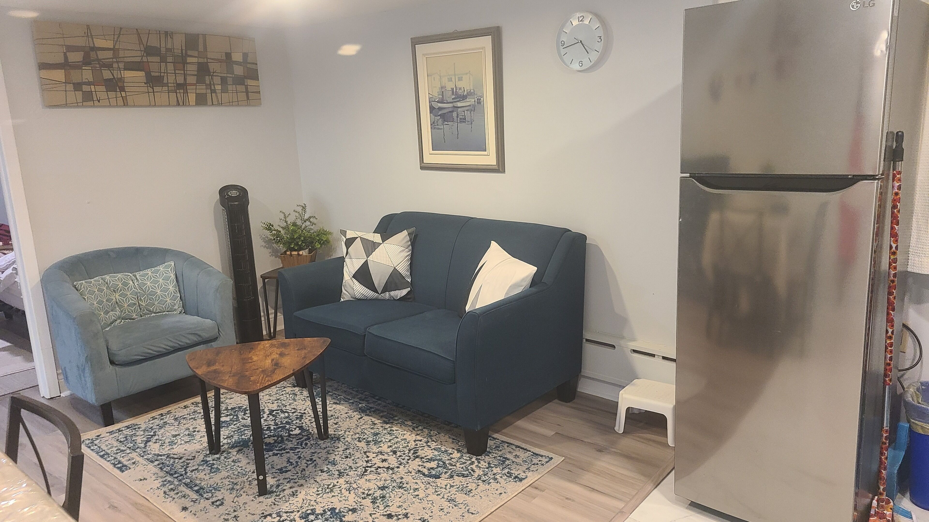 Una sala de estar con un sofá azul, una mesa de centro de madera, una guitarra, un cuadro enmarcado, un reloj y un refrigerador de acero inoxidable.