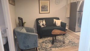 A living room with a sofa, a small table, a chair, a clock, and a refrigerator.