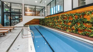 Une piscine intérieure dotée de bancs en bois, d’une échelle en métal et d’un mur décoré de feuillage verdoyant.