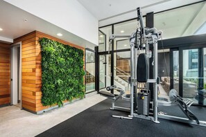 Ein Fitnessstudio mit Wandpflanze, Trainingsgeräten und Blick auf ein Gebäude durch große Fenster.