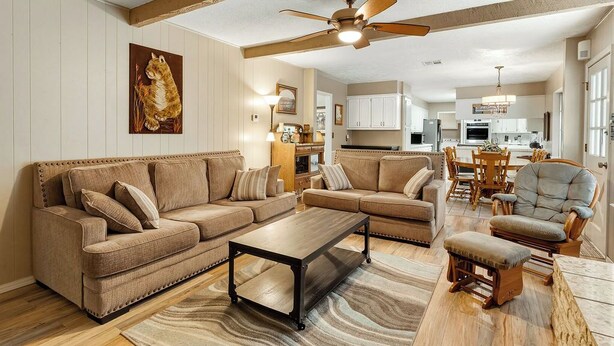 Una sala de estar con sofá, sillón y mesa de centro, un ventilador de techo y una cocina al fondo.