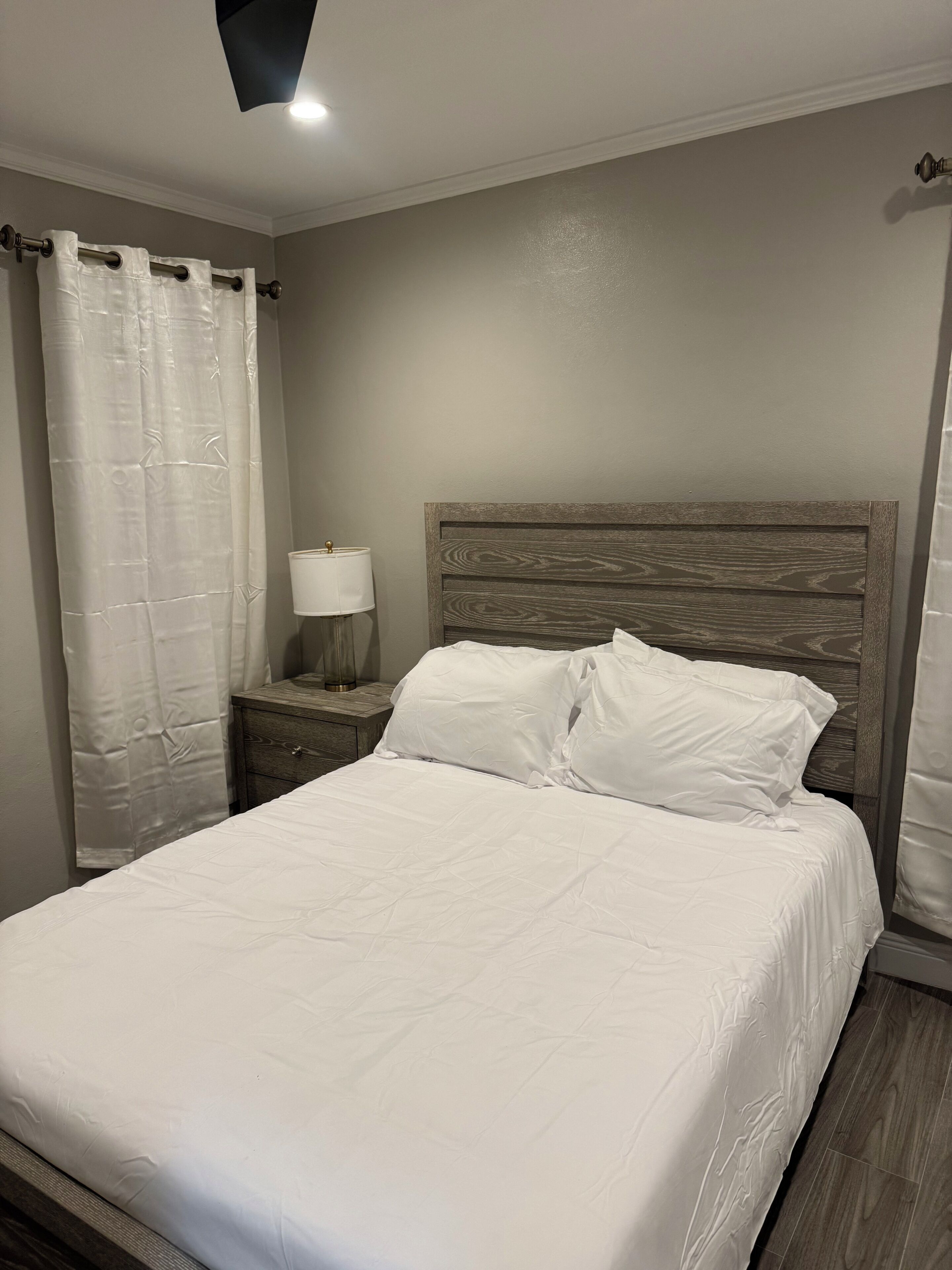 A bedroom with a large bed, a wooden headboard, a nightstand with a lamp, and a curtain.