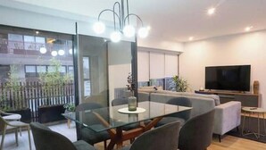 A modern living room with a glass dining table, grey sofas, a flat-screen TV, and a balcony with plants. - Apartamento Moderno em Edifício de Luxo - 3 beds/3 baths (Miraflores)