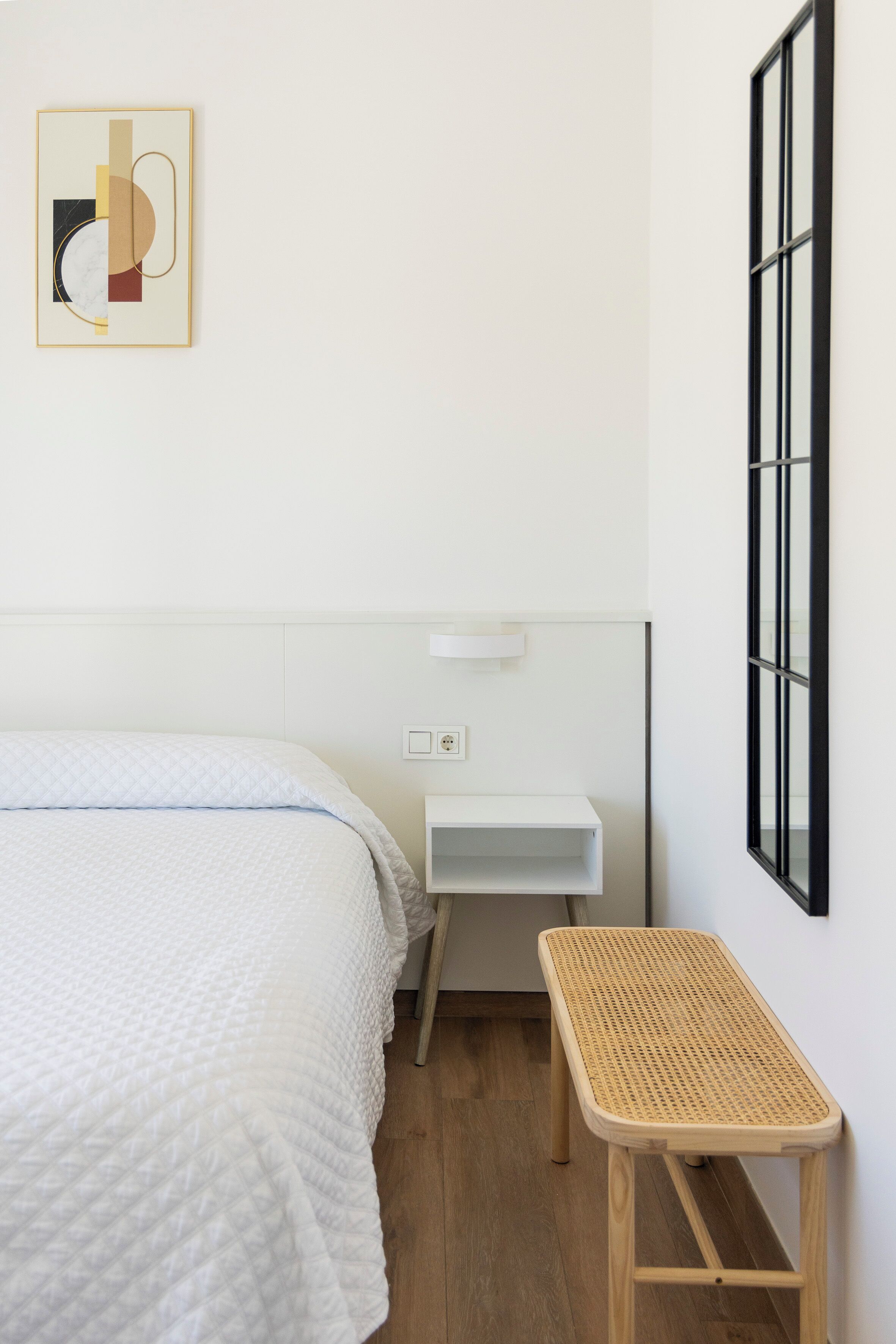 A minimalist room with a bed, a small white nightstand, a wicker side table, and a framed abstract artwork on the wall.