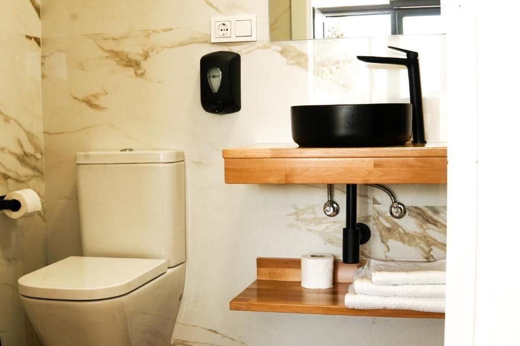 Un baño con inodoro, pared de mármol, dispensador de jabón negro, repisa de madera con toallas y lavabo negro.
