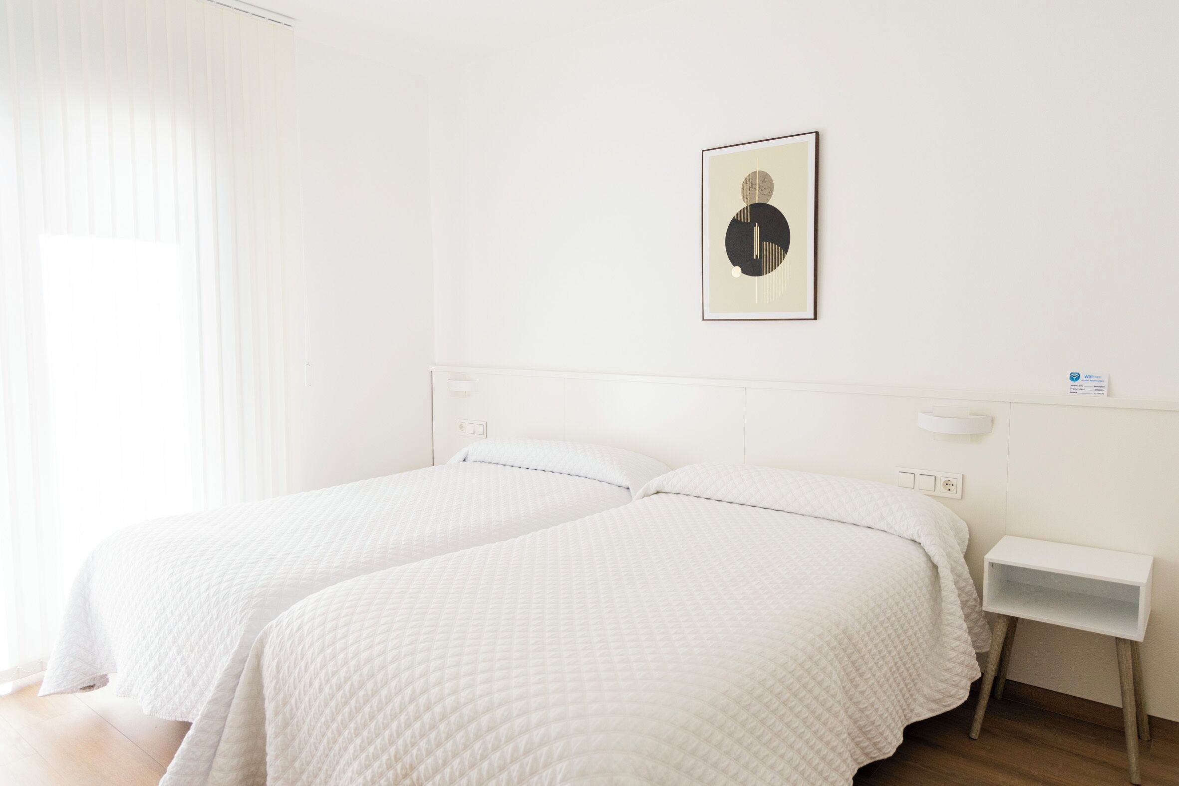 A room with two beds, a bedside table, a wall-mounted light, and a framed picture.