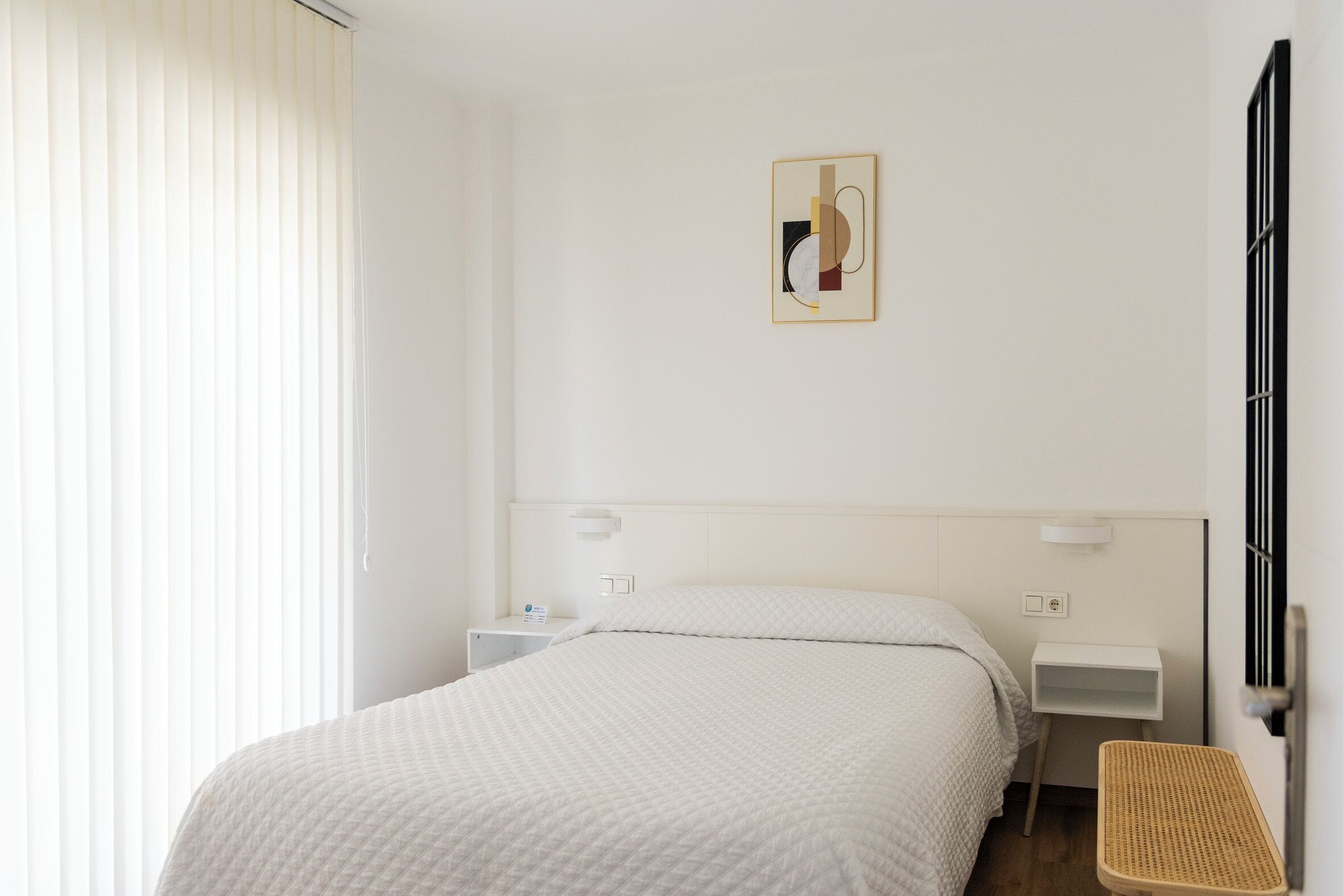 A hotel room with a bed, bedside tables, a wall-mounted artwork, and a window with blinds.