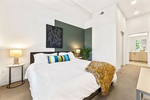 A hotel room with a bed, bedside tables, a lamp, a painting, and a view into the kitchen area.
