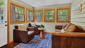 A sunroom with large windows, a wooden ceiling, a brown leather sofa, a round coffee table, and a patterned rug. -  Mountain lodge w/ cozy fall vibes! Slps 10, Wooded views, Hot tub & fire pit (McGaheysville)