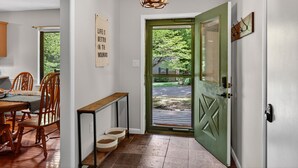A hallway with a wooden dining table, chairs, a bench, a chandelier, a green door, and a view of greenery outside. -  Mountain lodge w/ cozy fall vibes! Slps 10, Wooded views, Hot tub & fire pit (McGaheysville)