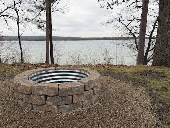 A circular stone fire pit surrounded by gravel, with a lake and trees in the background. - "West View Cabins" on Ostego Lake - Cabin #1 - Waterfront and on Trail 7 (Gaylord)