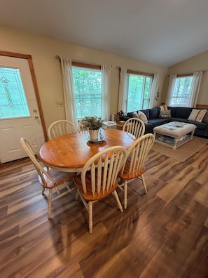 A dining area with a wooden table and chairs, a sofa, and a window with curtains. - "West View Cabins" on Ostego Lake - Cabin #1 - Waterfront and on Trail 7 (Gaylord)