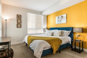 A bedroom with a bed, bedside table, lamp, and artwork on the wall.
