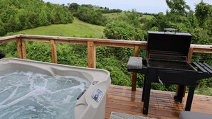 Un jacuzzi y una parrilla en una terraza de madera con vistas a un paisaje verde.