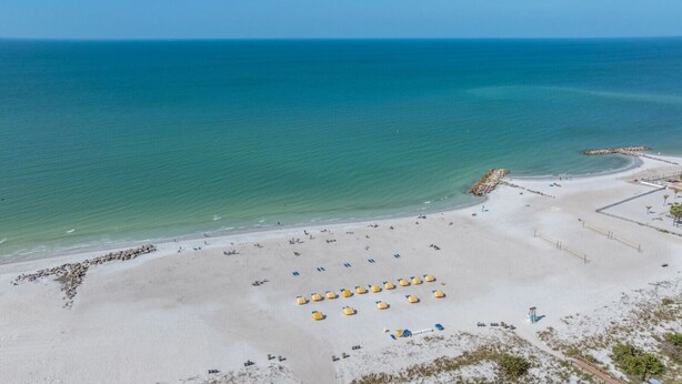 Vista aérea de uma praia com céu claro, mar calmo e faixa de areia com guarda-sóis e um paredão rochoso.