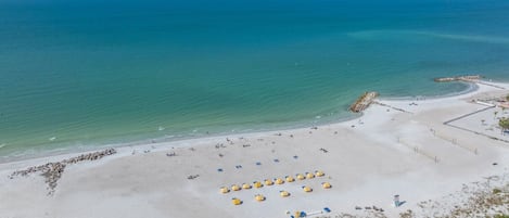 Vista aérea de una playa con cielo despejado, mar tranquilo y una orilla arenosa con sombrillas y un rompeolas rocoso.
