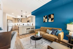 A modern living room with a sofa, coffee table, and a kitchen area with a refrigerator and bar stools.