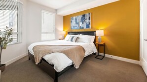 A bedroom with a bed, two bedside lamps, a wall art piece, a potted plant, and a window with blinds. - Chic & Cozy Condo | South Coast | Mins to Irvine (Santa Ana)