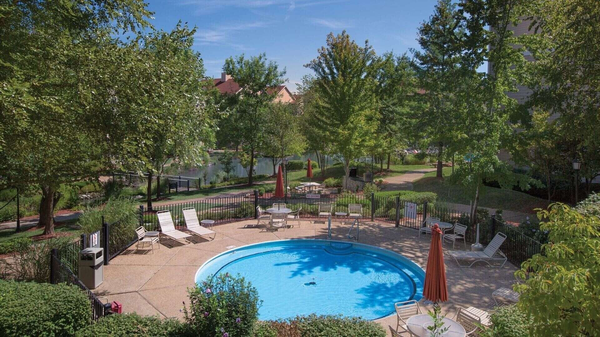 A swimming pool surrounded by lounge chairs and umbrellas, with a fence and trees in the background.