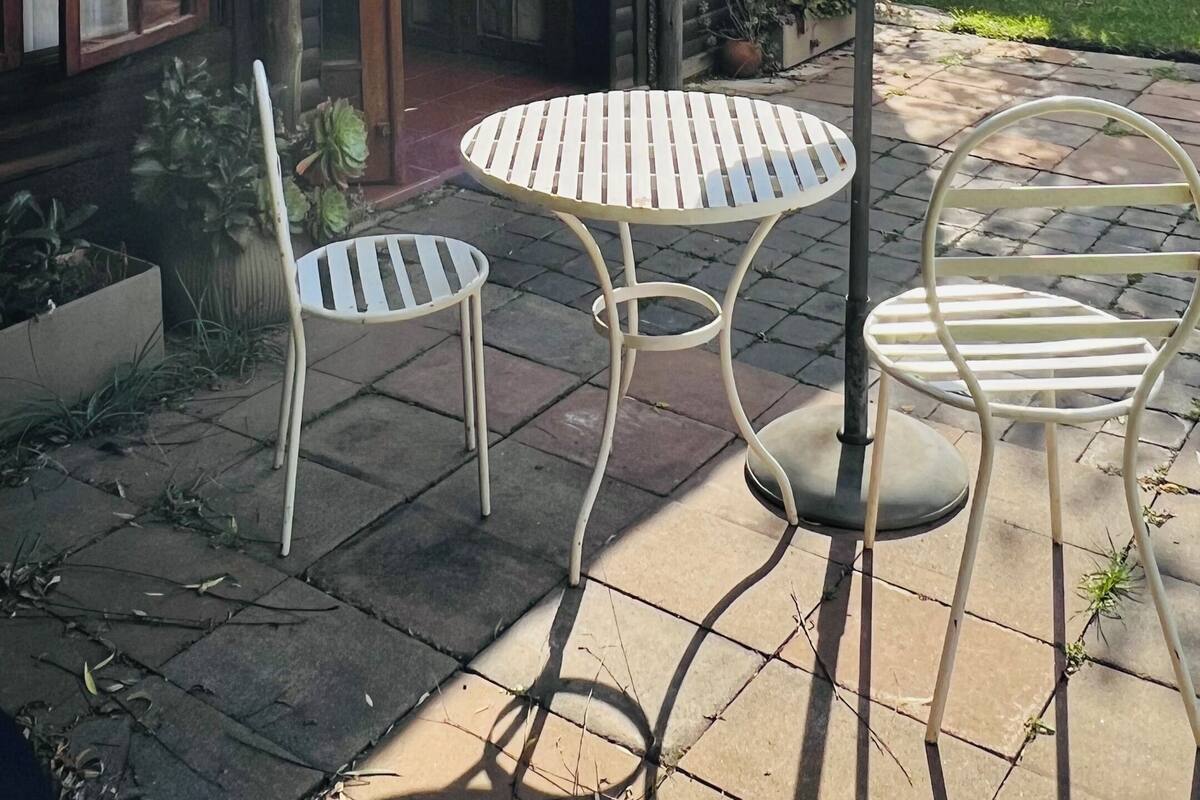 Een landelijk gebouw met rieten dak, een houten deur en ramen. Er staat een ronde tafel met een witte parasol, twee witte stoelen en een bloempot.