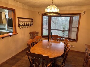Ein Esszimmer mit einem runden Holztisch, sechs Stühlen, einer Chandelierleuchtre, einem Fenster mit Jalousien sowie einer Küchenzeile mit Kühlschrank und Geschirrständer.