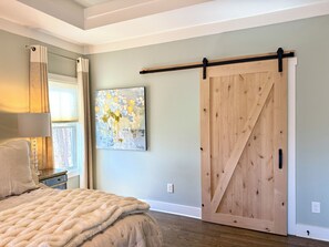 A bedroom with a bed, a painting, a sliding barn door, and a window with curtains. - The Painted Fox - Beautiful Big Canoe, GA - nestled in the Blue Ridge Mountains! (Jasper)