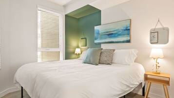A bedroom with a large bed, a nightstand with a lamp, a painting on the wall, and a window with blinds.
